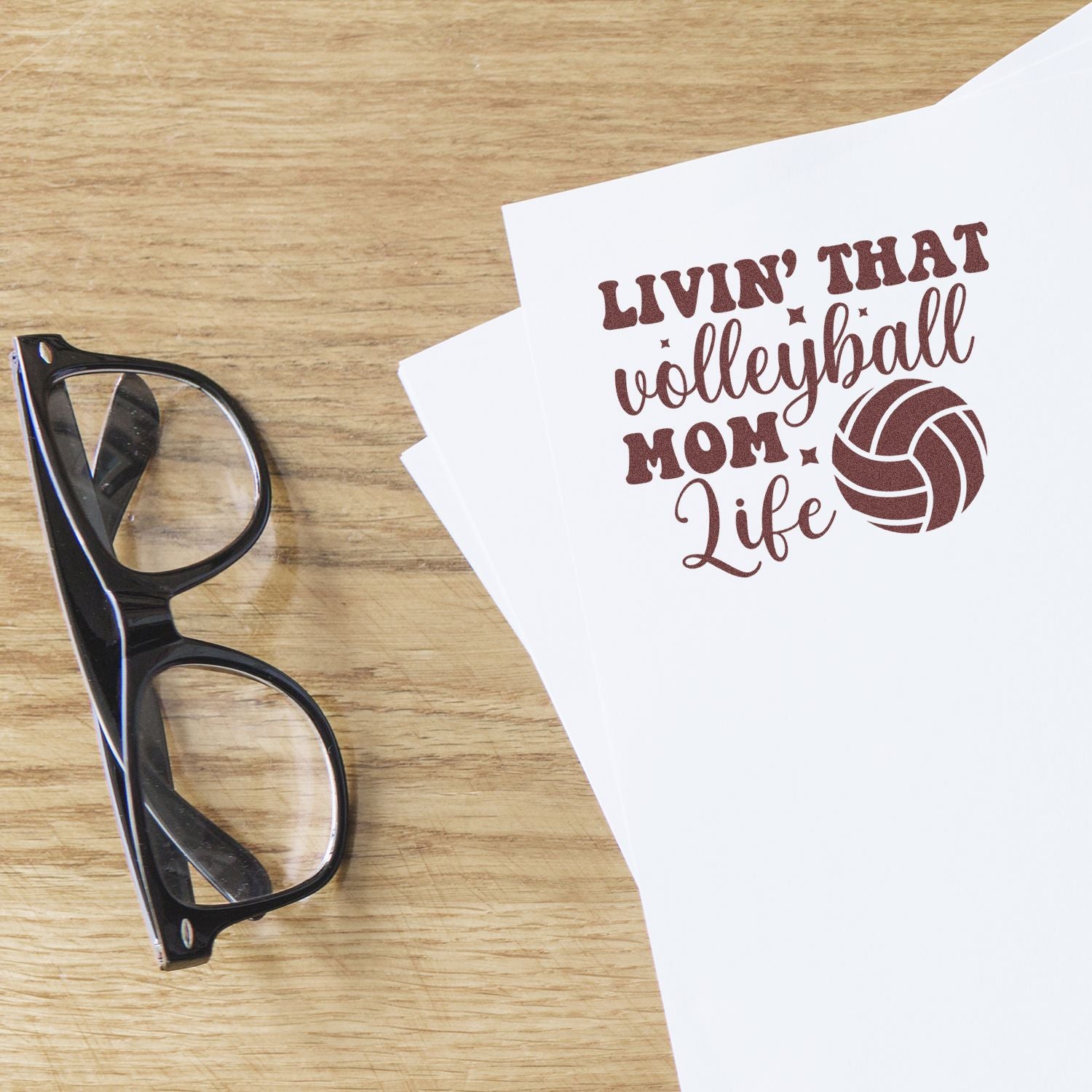 Brown volleyball mom stamp seal on white paper beside black glasses on a wood table