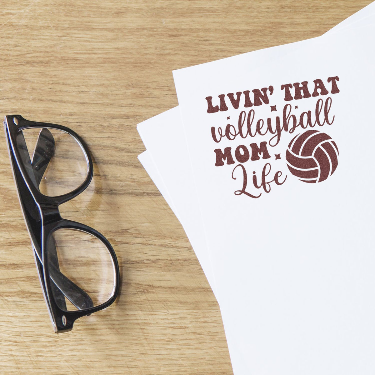 Brown volleyball mom life stamp design with volleyball icon on white paper, beside black eyeglasses on wood