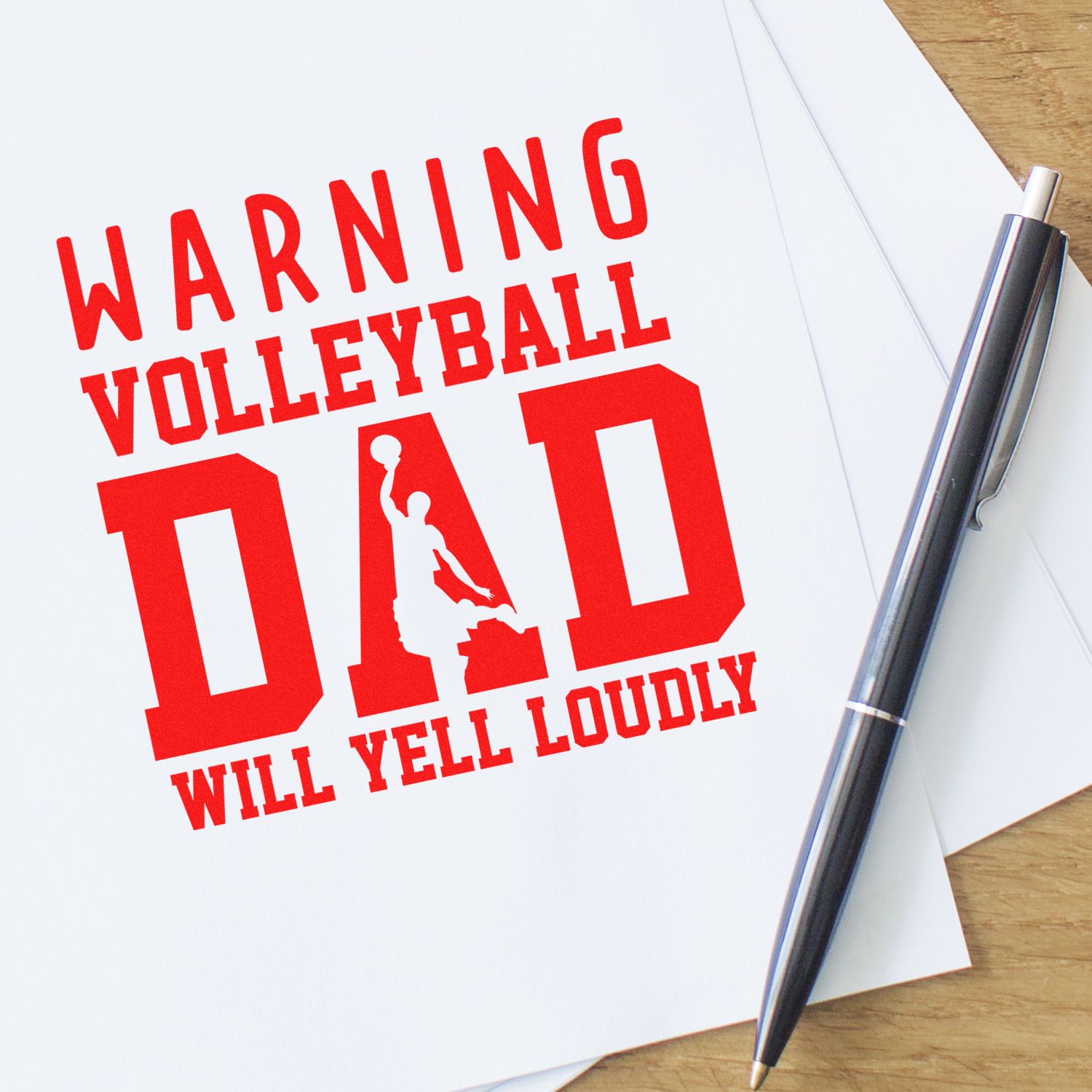 Red volleyball dad warning stamp and seal on white paper beside a silver pen on a wood desk.