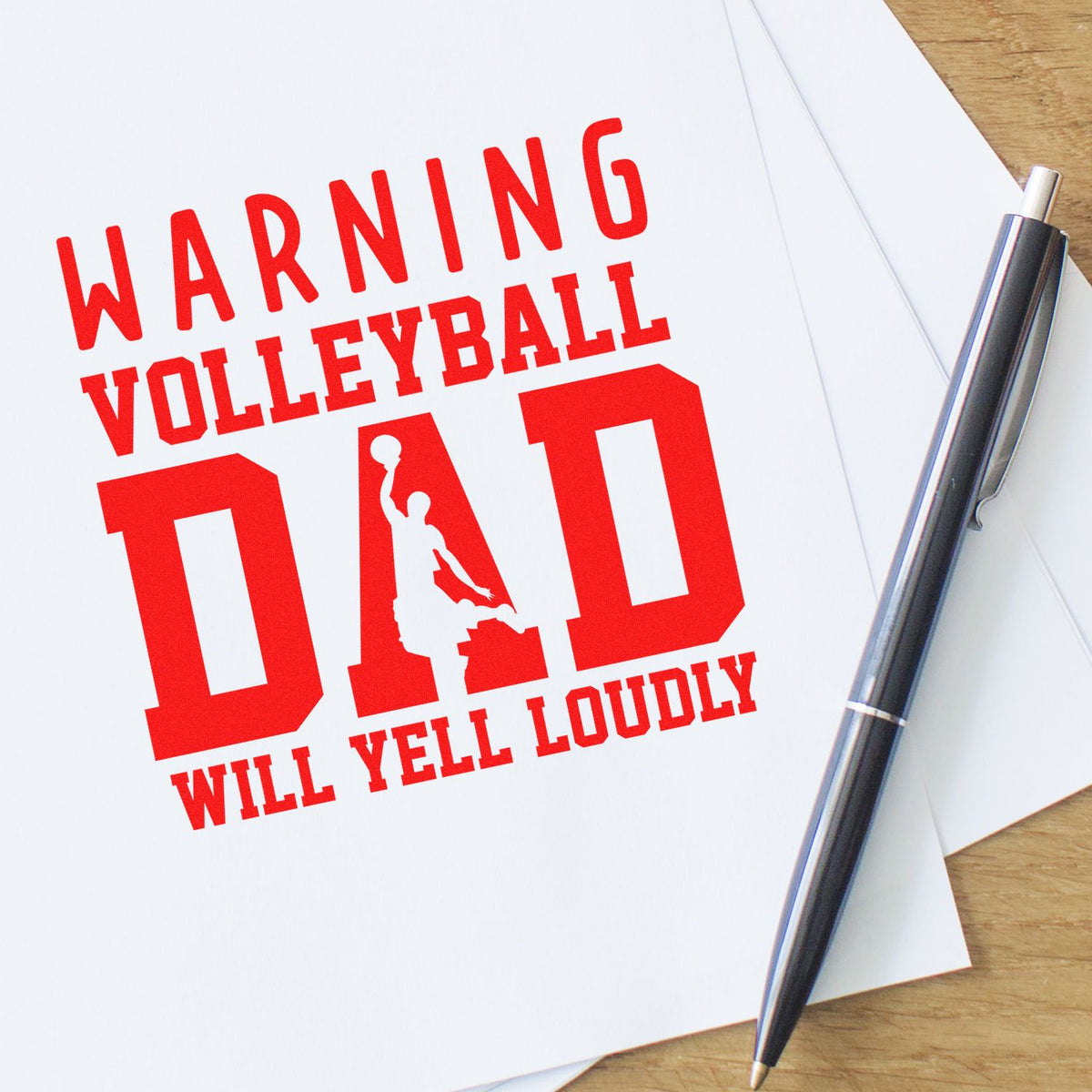 Red custom stamp design reading Warning Volleyball Dad Will Yell Loudly on white paper, with a silver pen.