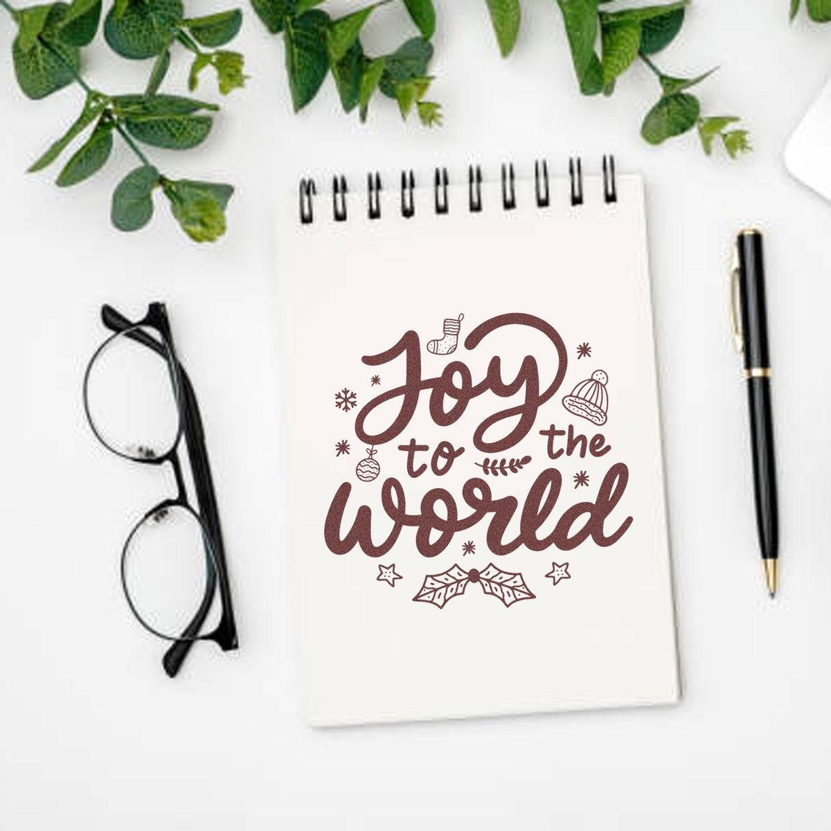 Brown holiday stamp seal reading Joy to the world on a white spiral notebook, with Christmas icons, glasses, pen, and greenery.