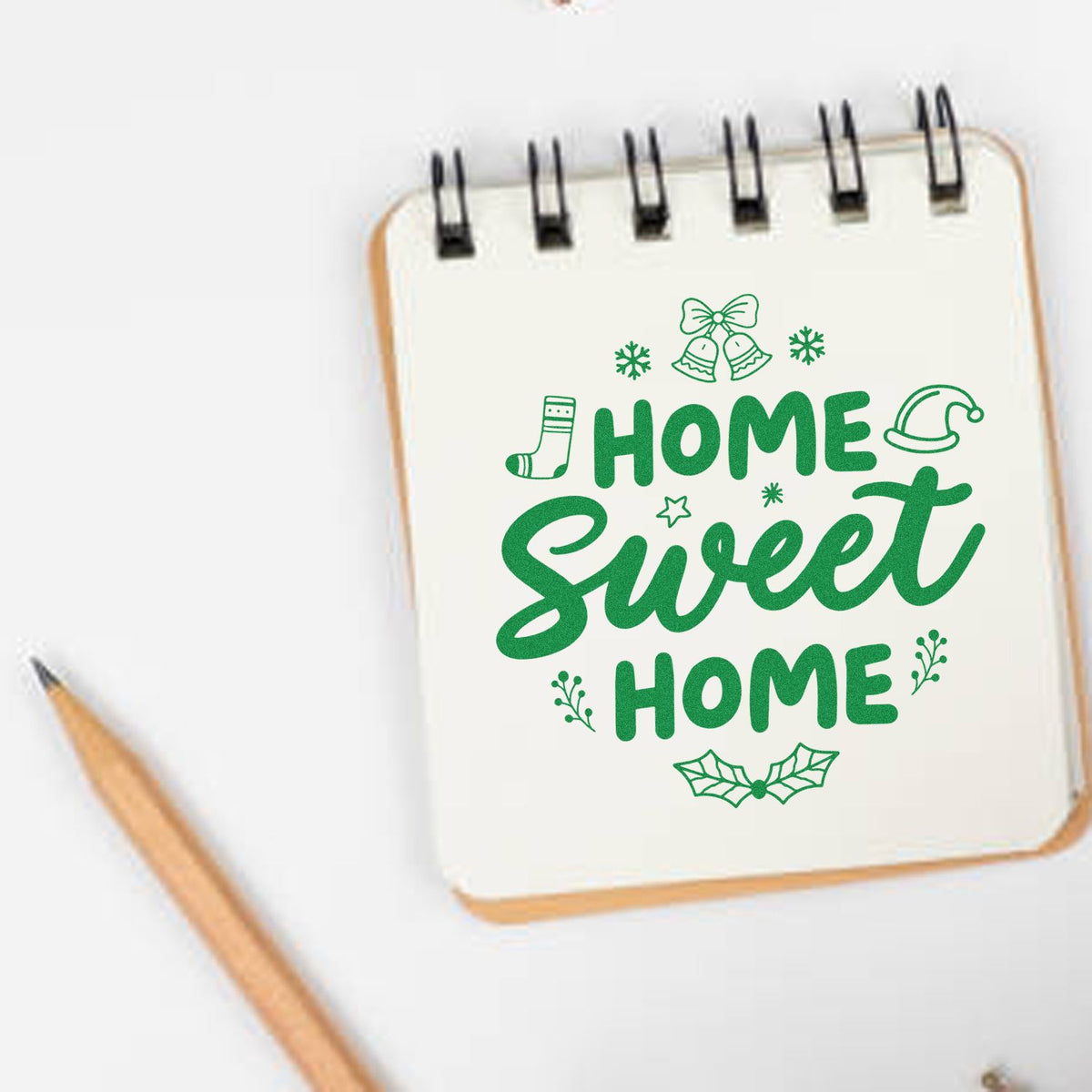 Green holiday stamp/seal design reading HOME Sweet HOME on a white notebook, with bells, holly, snowflakes and a pencil.