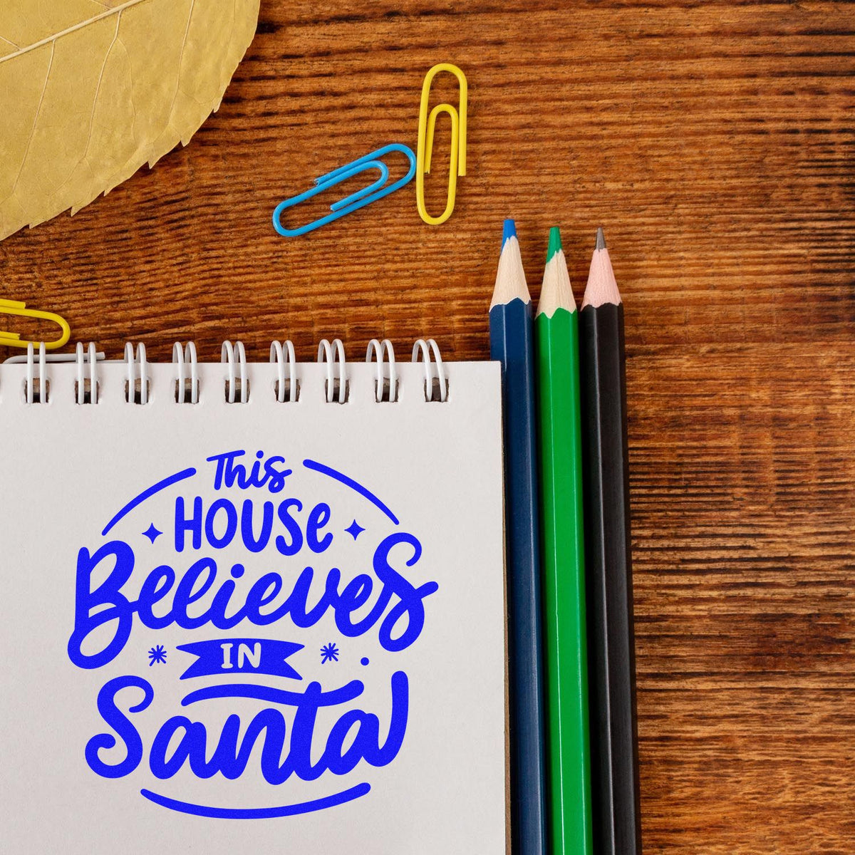 Blue This House Believes in Santa stamp/seal on a spiral notebook, with colored pencils and paper clips on a wooden desk.