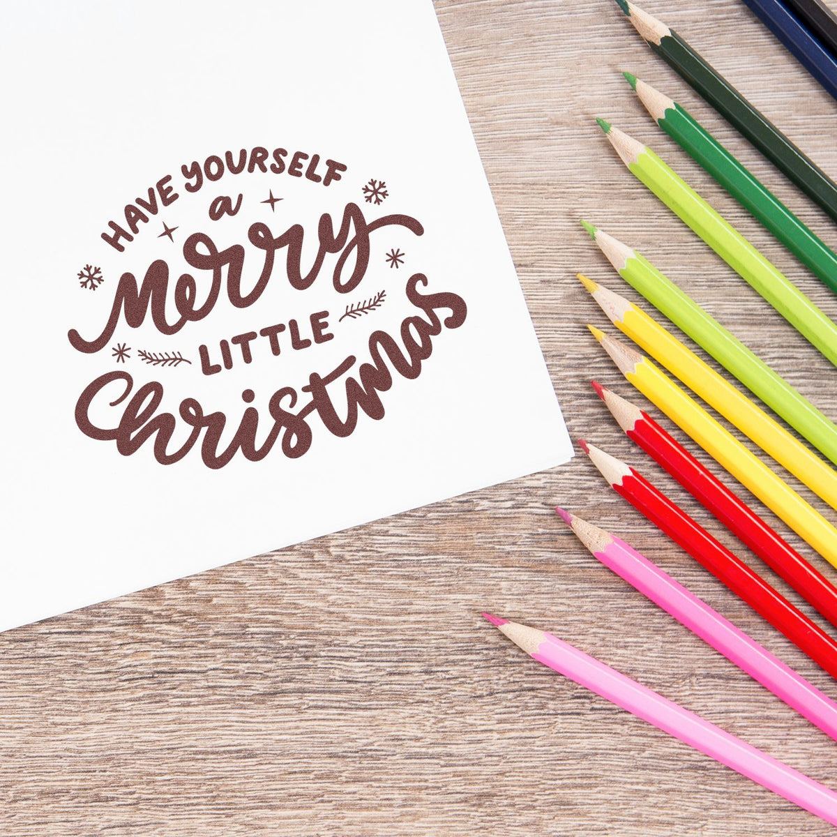 Brown Christmas stamp seal with Have Yourself a Merry Little Christmas beside colorful pencils on a wood desk.