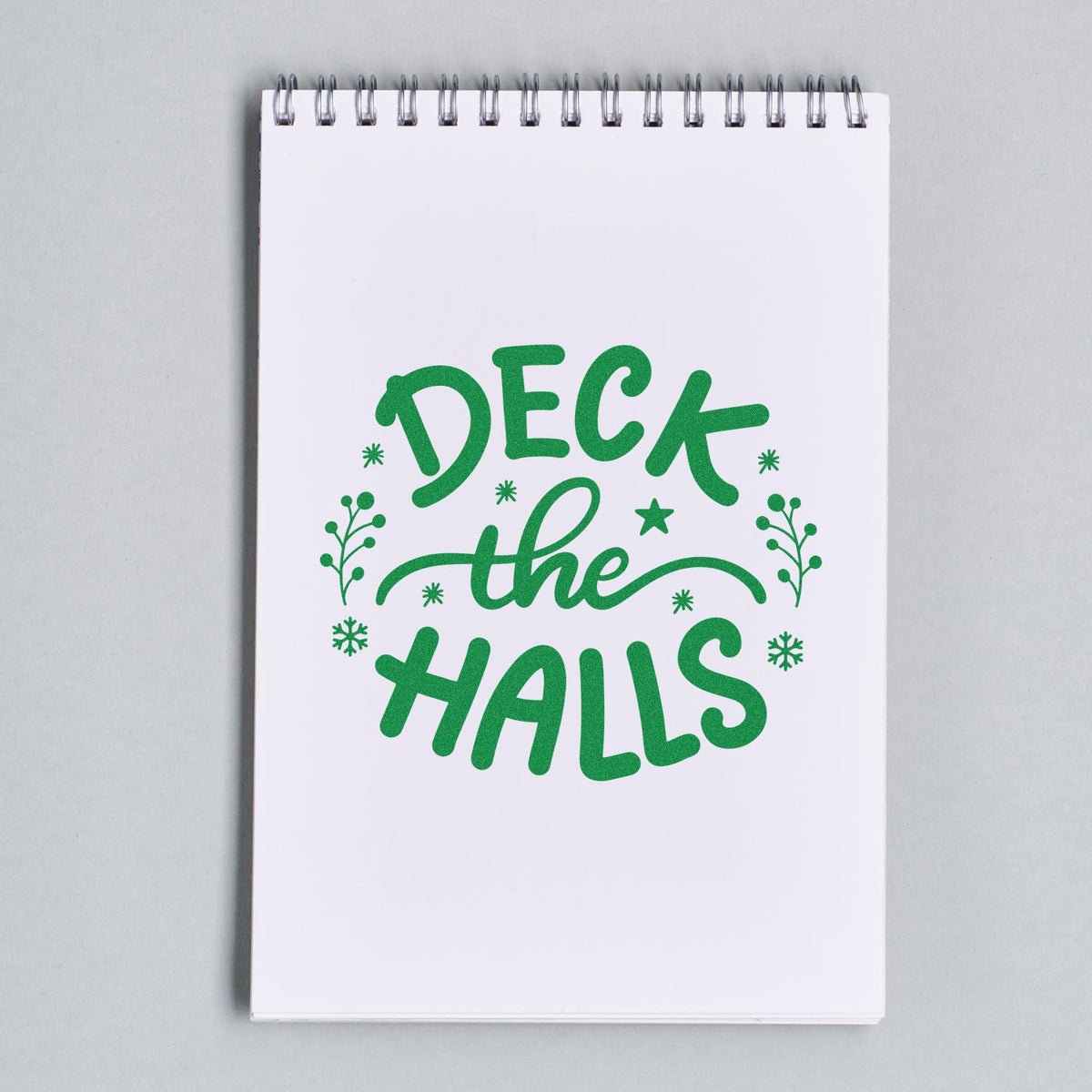 Green holiday stamp and seal design reading Deck the Halls with snowflakes and sprigs on a white spiral notebook page.