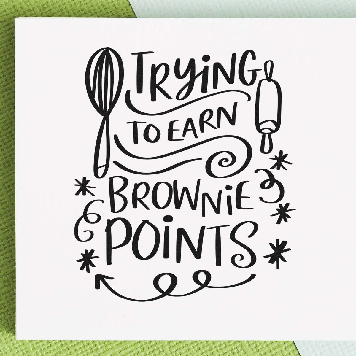 Black stamp and seal design with whisk and rolling pin, reading Trying to earn brownie points in black on white paper.