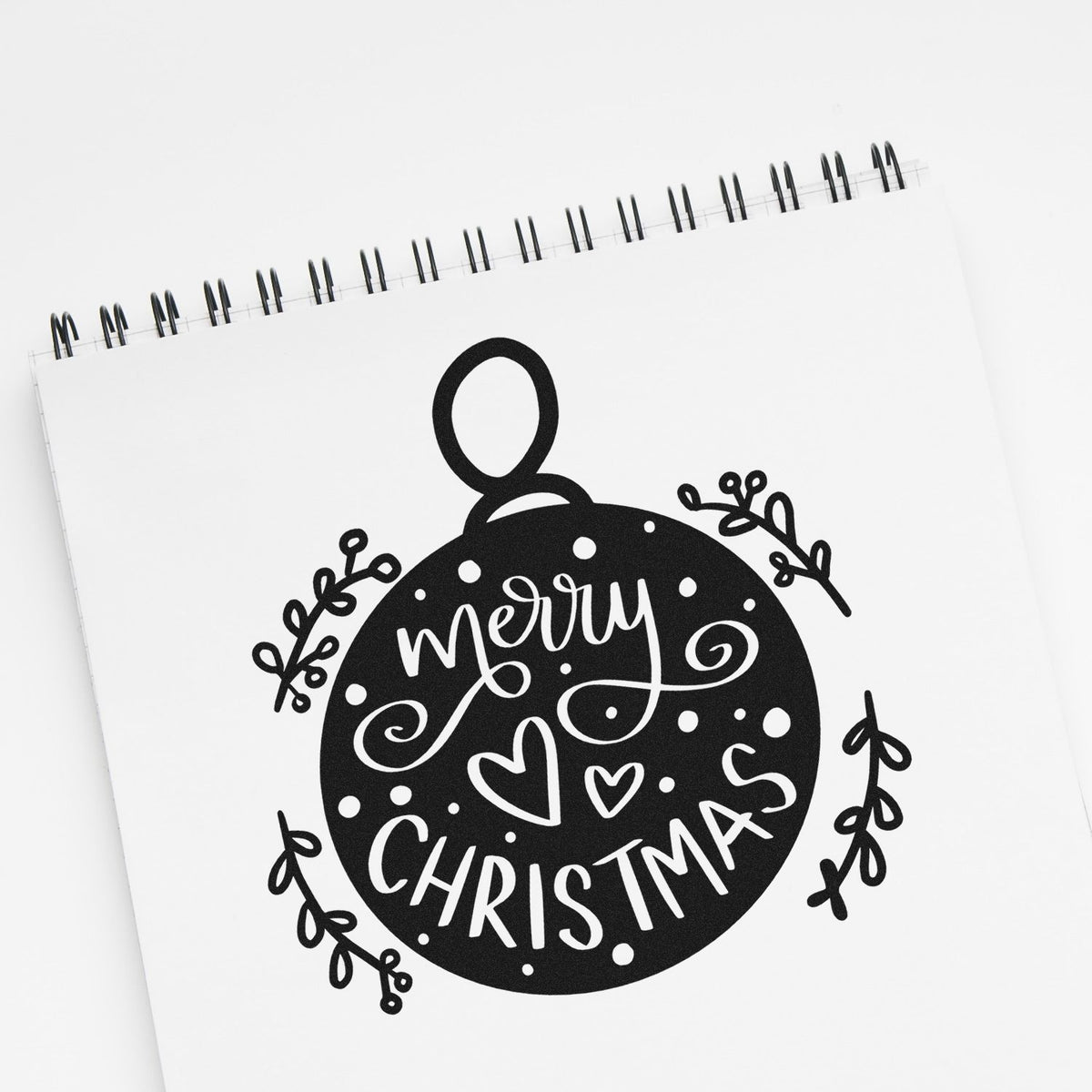 Black-and-white Merry Christmas stamp and seal design with a round ornament, hearts, and holly sprigs on a spiral notebook.