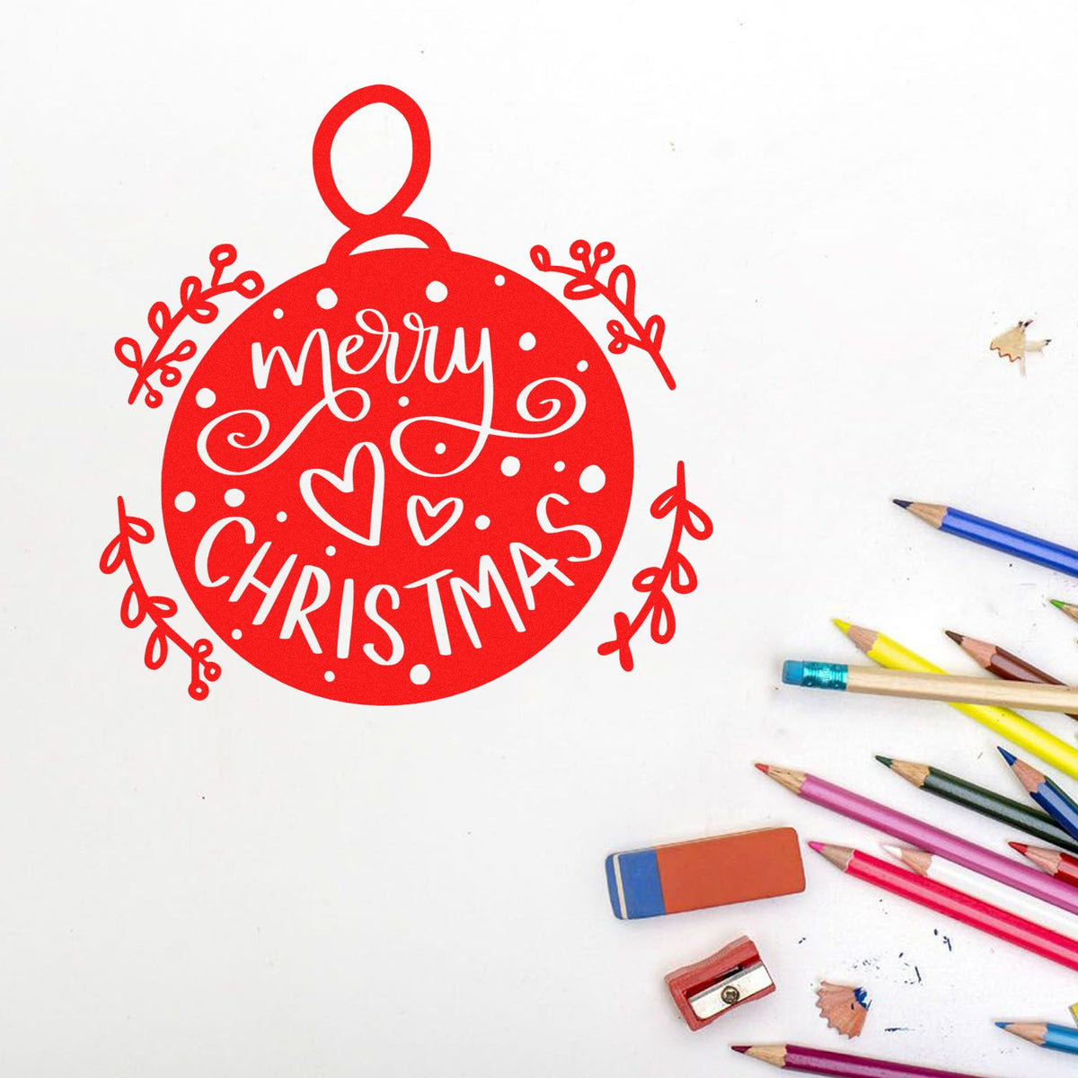 Red Merry Christmas ornament stamp and seal with white lettering, hearts and dots on a white background, beside colored pencils.