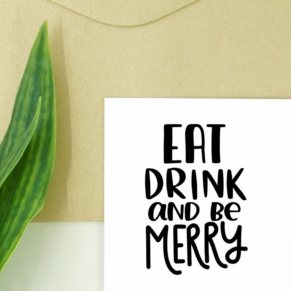 Black stamp and seal reading EAT DRINK and be MERRY on white card with beige background and green leaf.