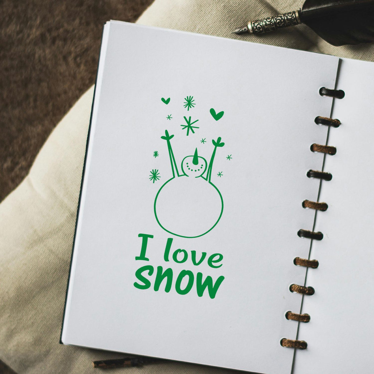 Green I love snow stamp with a snowman, hearts and stars on a white spiral-bound notebook page.