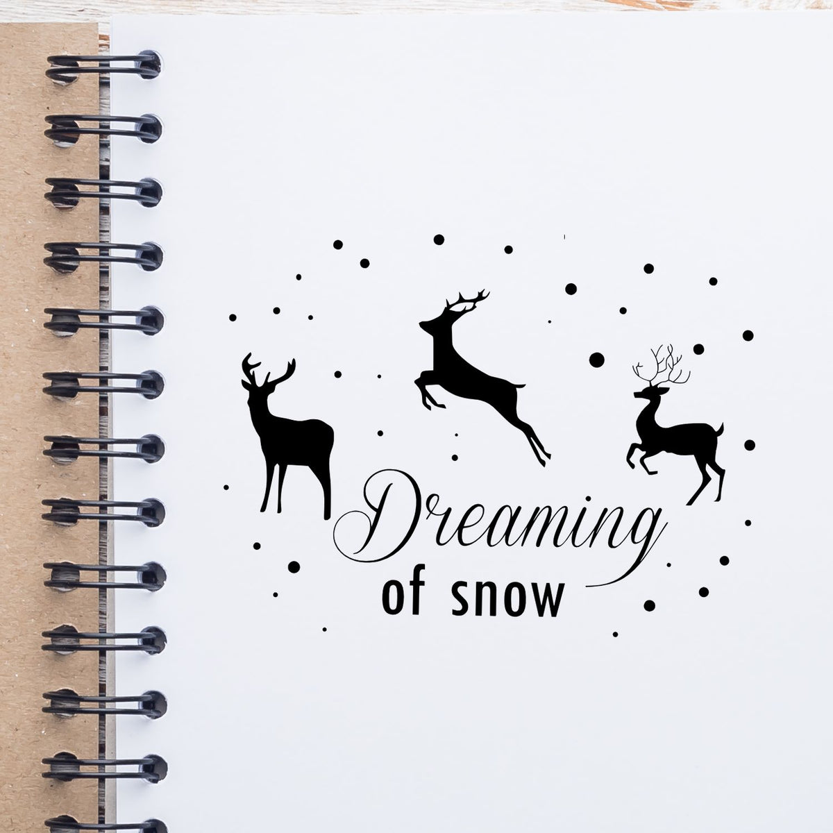 Black deer stamp seal reading Dreaming of snow on white spiral notebook, with black dots and kraft cover.