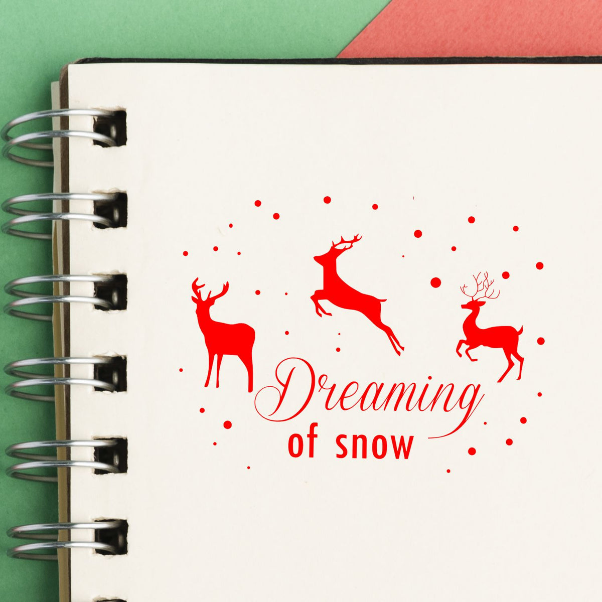 Red deer stamp and seal design with Dreaming of Snow text on cream spiral notebook page, red accents, green and red background.