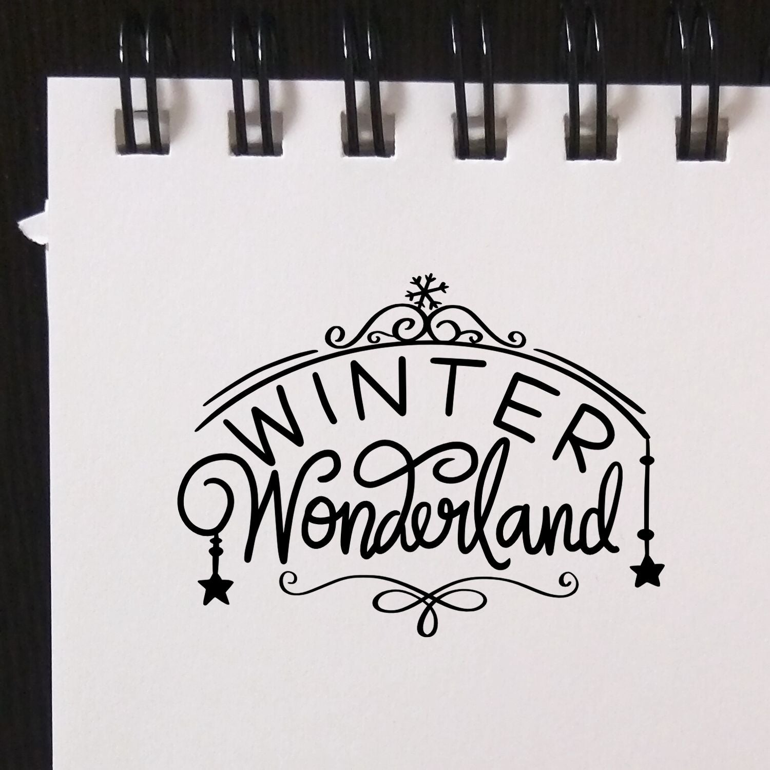 Black winter wonderland stamp and seal design in black on white spiral-bound paper, with snowflake, stars, and ornate swirls.