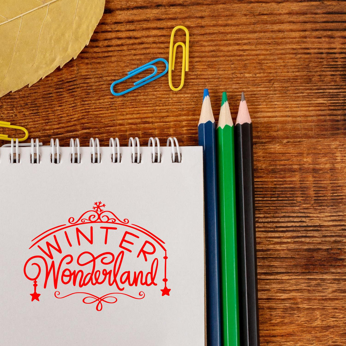Red Winter Wonderland decorative stamp on white spiral notebook, with blue, green, and black pencils on a wood desk.