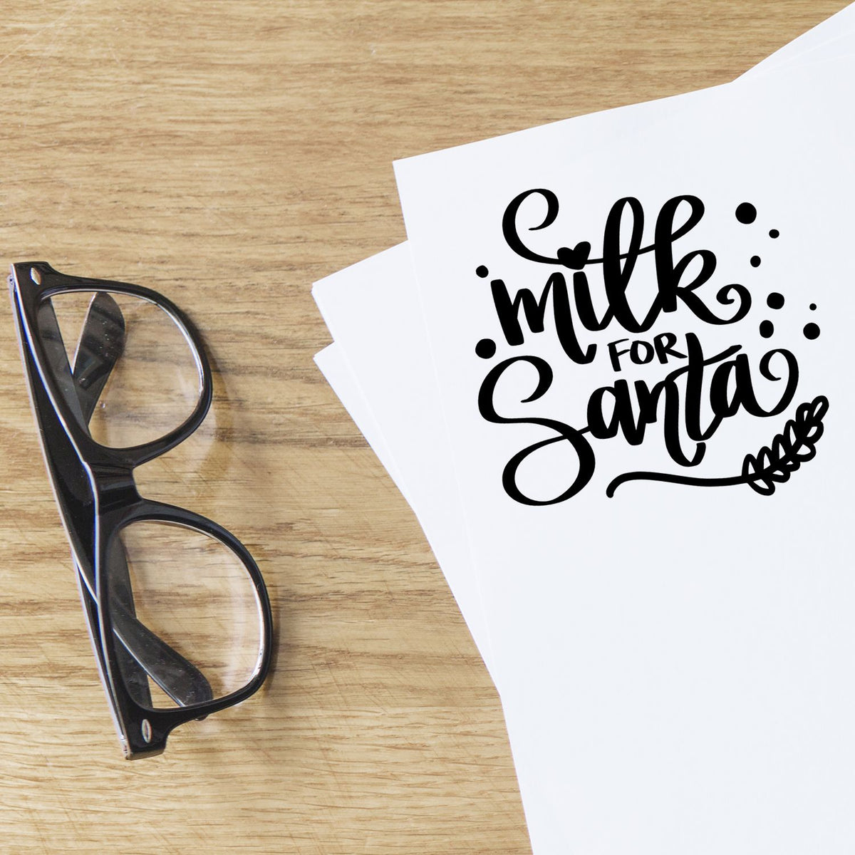 Black stamp and seal design reading milk for Santa on white paper, beside black eyeglasses on a wood table.