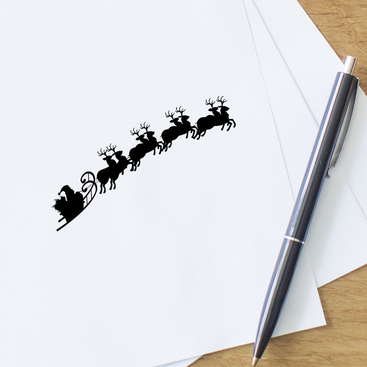 Black Santa sleigh and reindeer stamp on white paper beside a silver pen, showing a custom stamp and seal design.