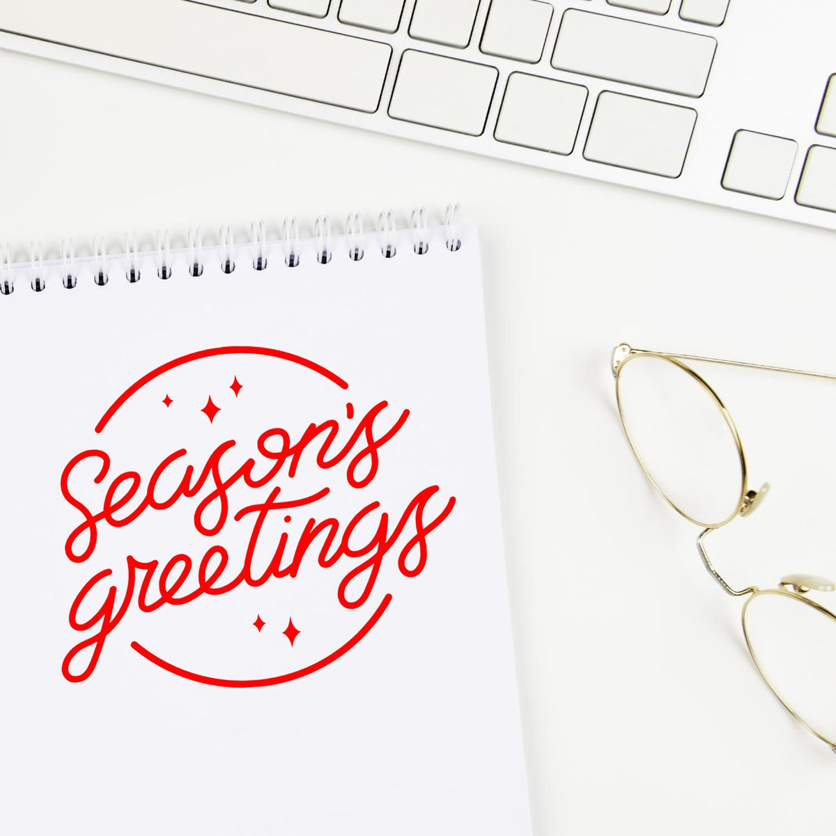 Red Seasons greetings stamp and seal on a white notepad, beside gold glasses and a white keyboard.