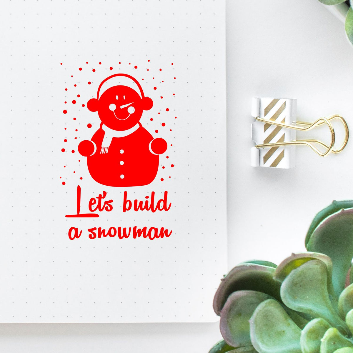Red snowman rubber stamp with Lets build a snowman on a white dotted notebook page, clipped with a gold binder clip.