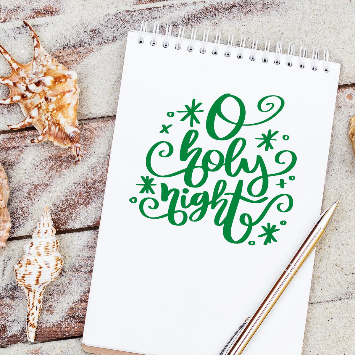 Green Oh Holy Night stamp and seal design on white spiral notebook, with seashells and pen on sandy wood background.