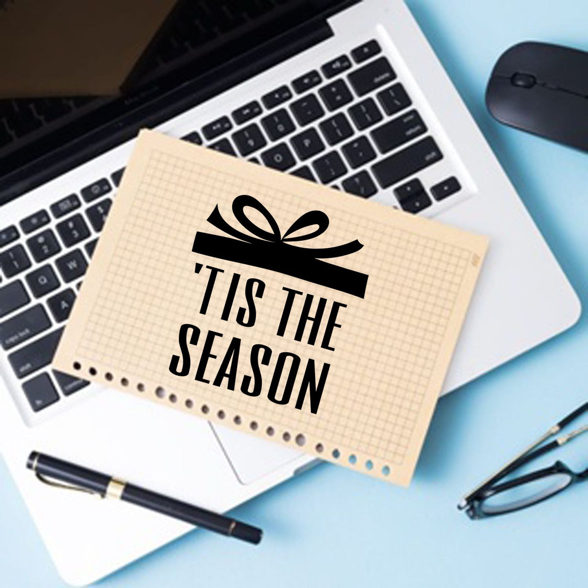 Beige holiday stamp and seal card with black gift icon and Tis the Season text on a laptop, blue desk, pen, mouse and glasses.