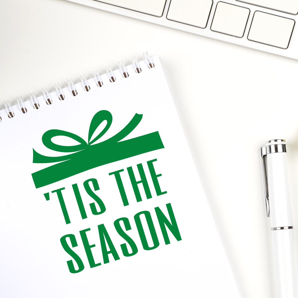 Custom stamp and seal product with green Tis the Season gift ribbon on a white spiral notebook, beside a white pen.
