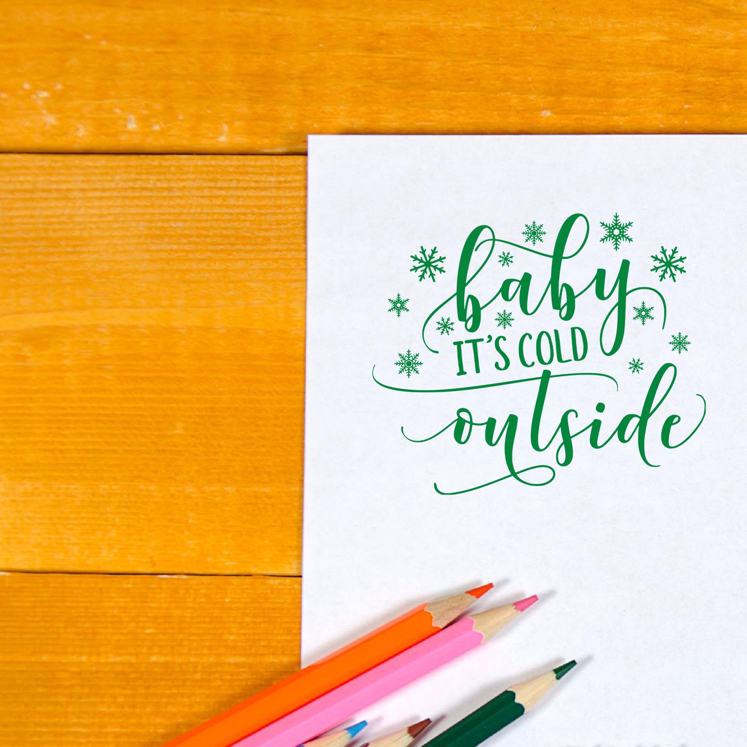 Green baby its cold outside stamp and seal design with snowflakes on white paper, beside orange, pink, and green pencils.