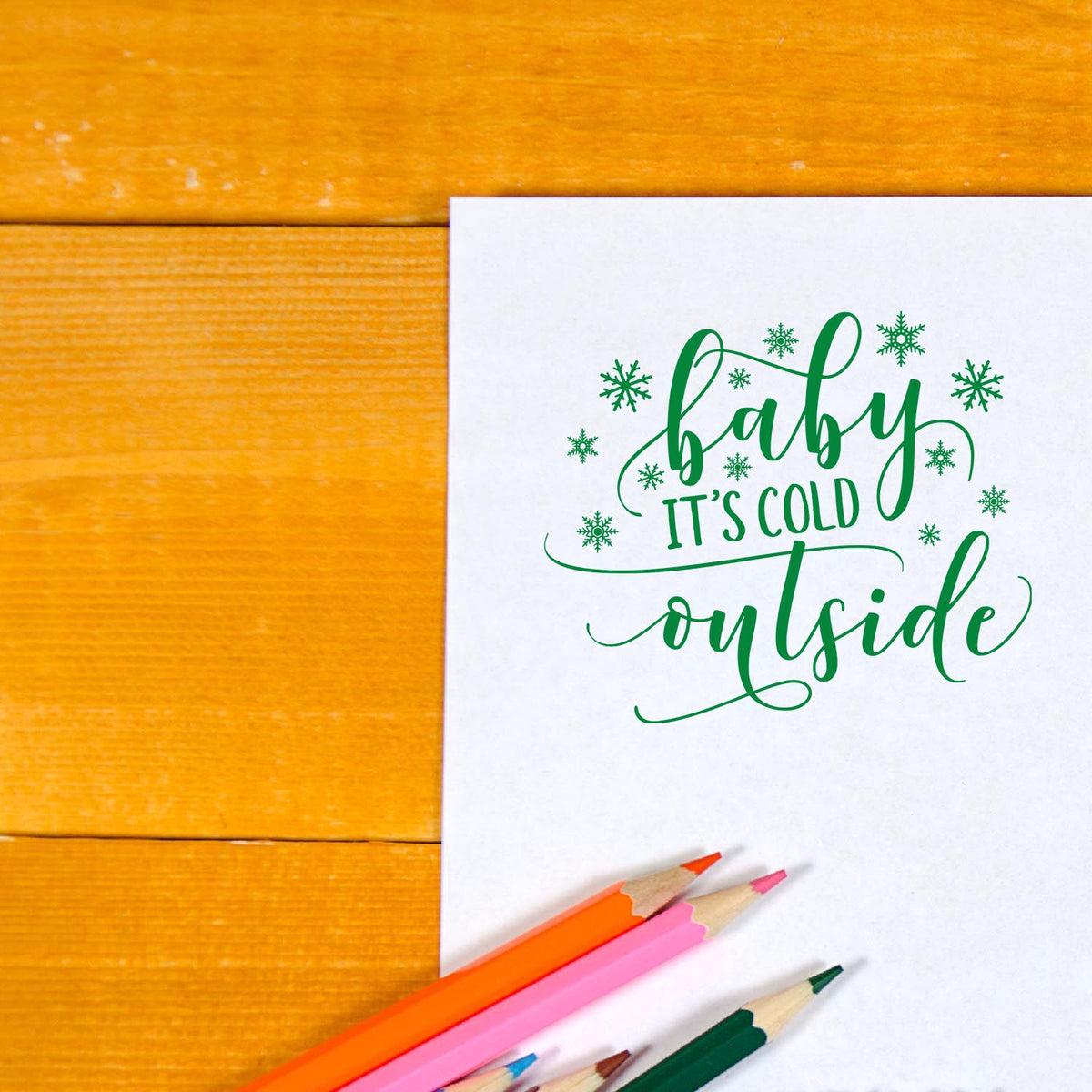 Green holiday stamp design with snowflakes reading baby its cold outside on white paper, beside colorful pencils on wood.