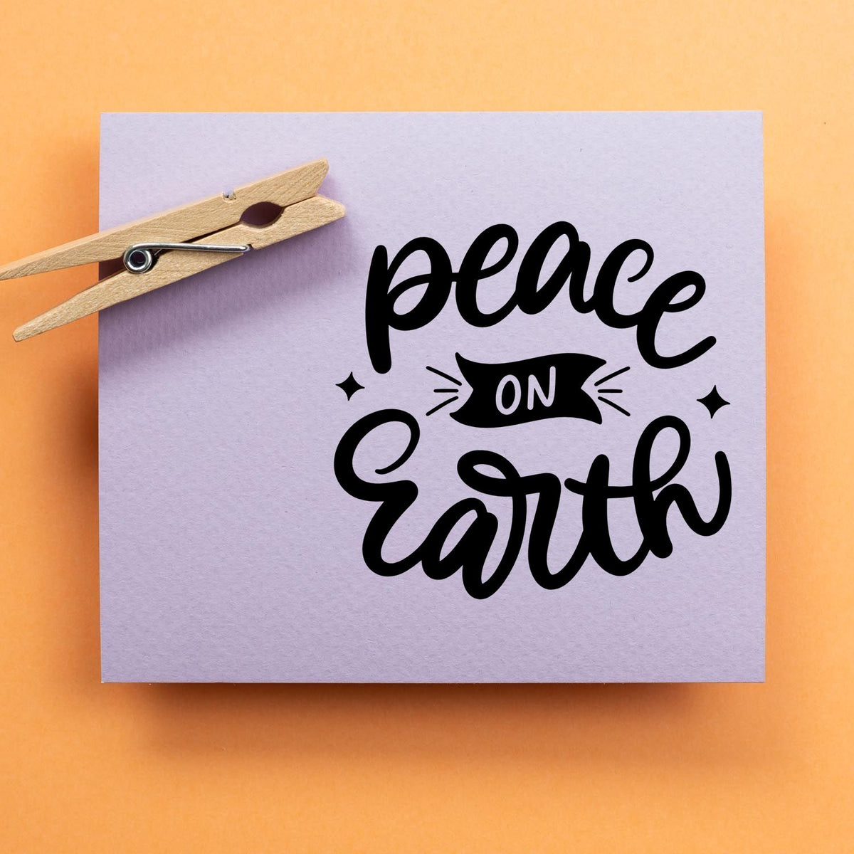Wooden clothespin-style stamp and seal on lavender paper, with black peace on Earth lettering on orange background.