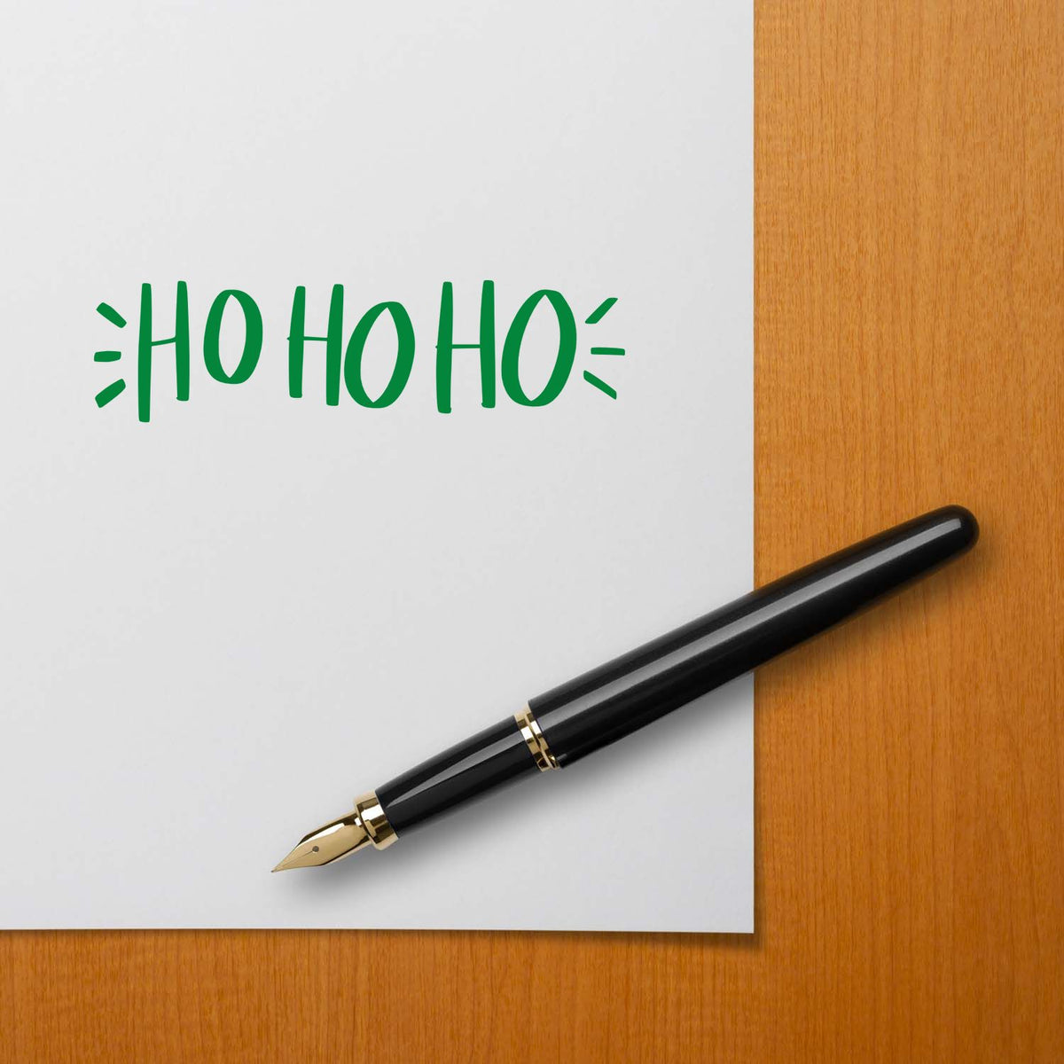 Green HO HO HO stamp design on white paper with a black fountain pen on a warm wood desk.