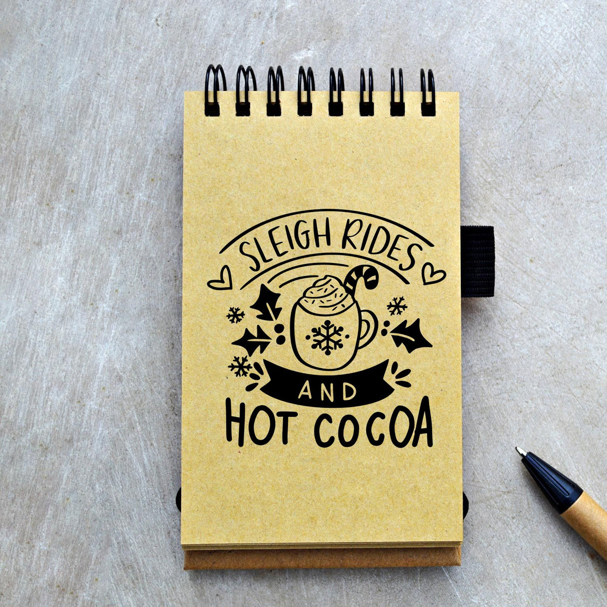 Kraft spiral notepad with black holiday stamp design: Sleigh Rides and Hot Cocoa, featuring a cocoa mug, snowflakes, and pen.