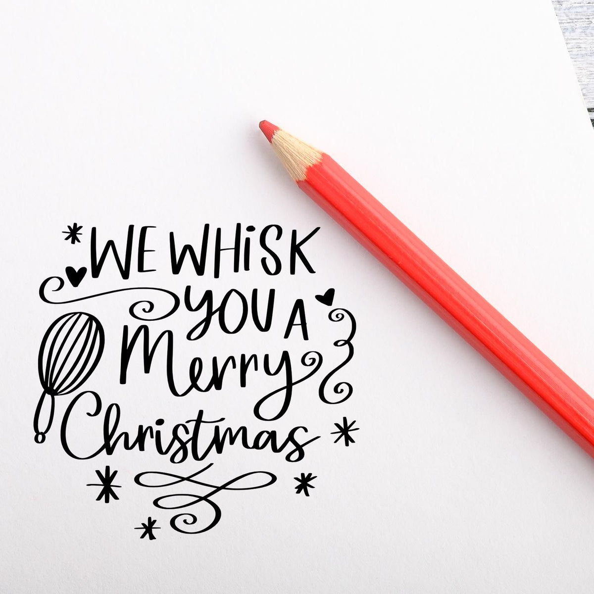 Decorative Christmas stamp and seal design in black, reading We wish you a Merry Christmas, with a red pencil on white paper.