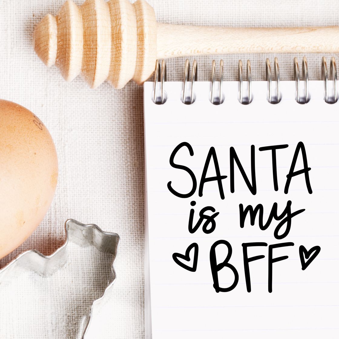 Wooden stamp and metal seal cutter beside a white notebook reading Santa is my BFF in black on cream fabric.