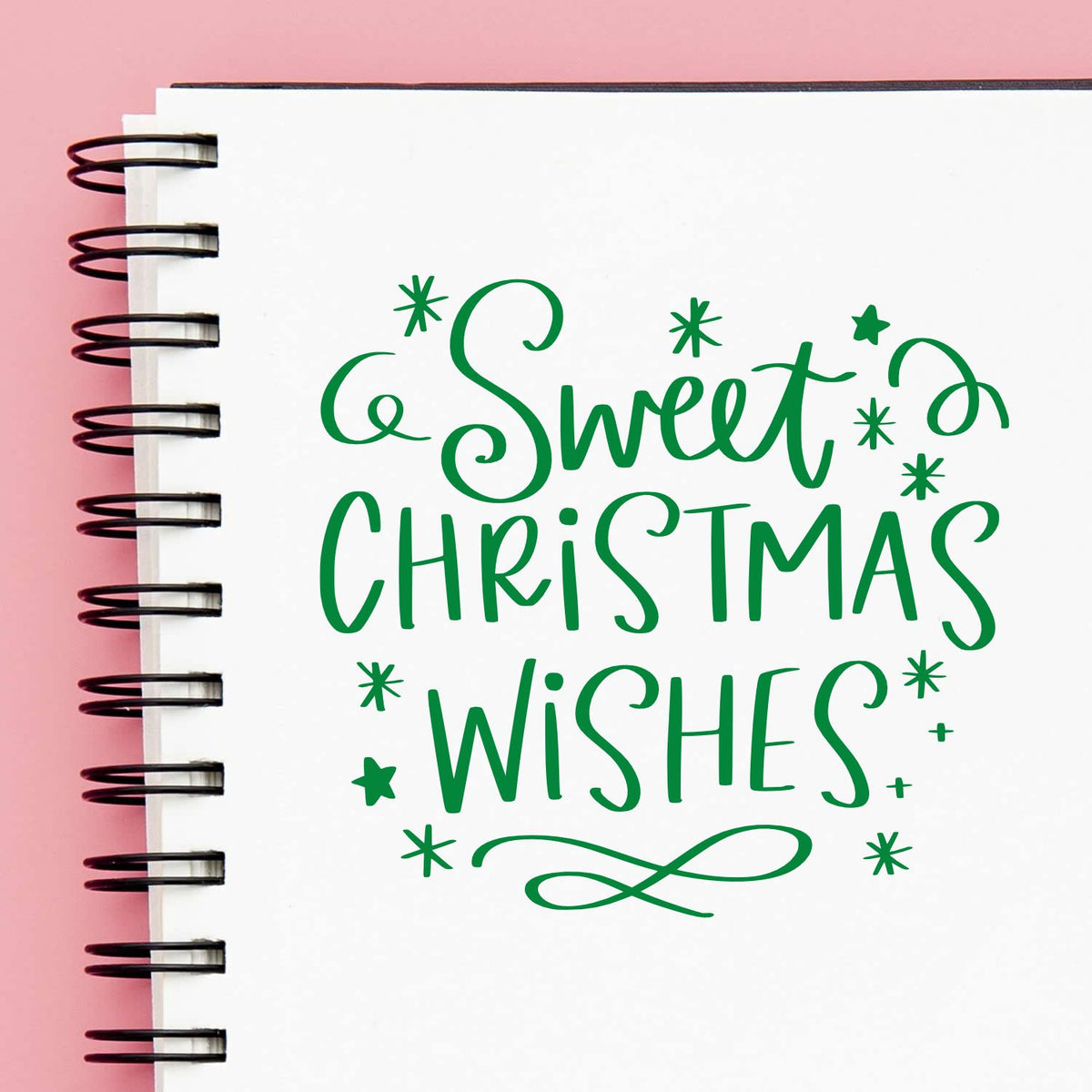 Green Sweet Christmas Wishes stamp design on white spiral notebook with pink background and black coil binding.