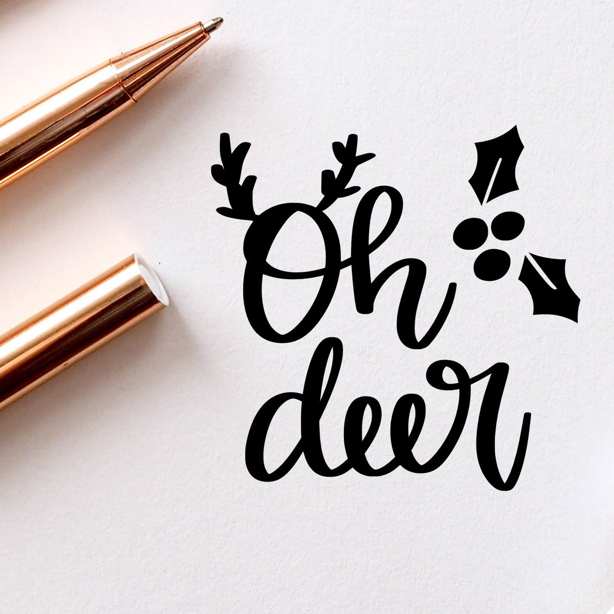 Black Oh deer holiday stamp and wax seal design with antlers, holly berries, and rose gold stamp tools on white paper.