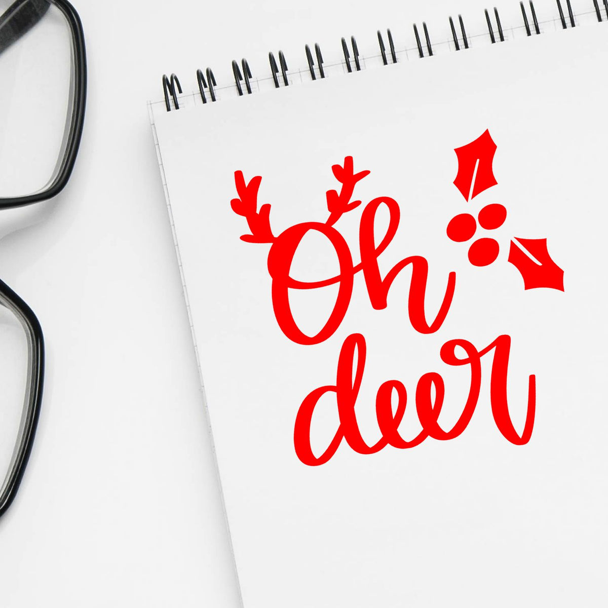 Red holiday stamp seal reading Oh deer on a white spiral notebook, with eyeglasses at left.