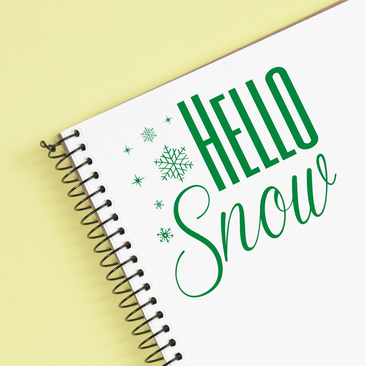 Green HELLO Snow stamp and seal design on a white spiral notebook with snowflakes, set against a pale yellow background.