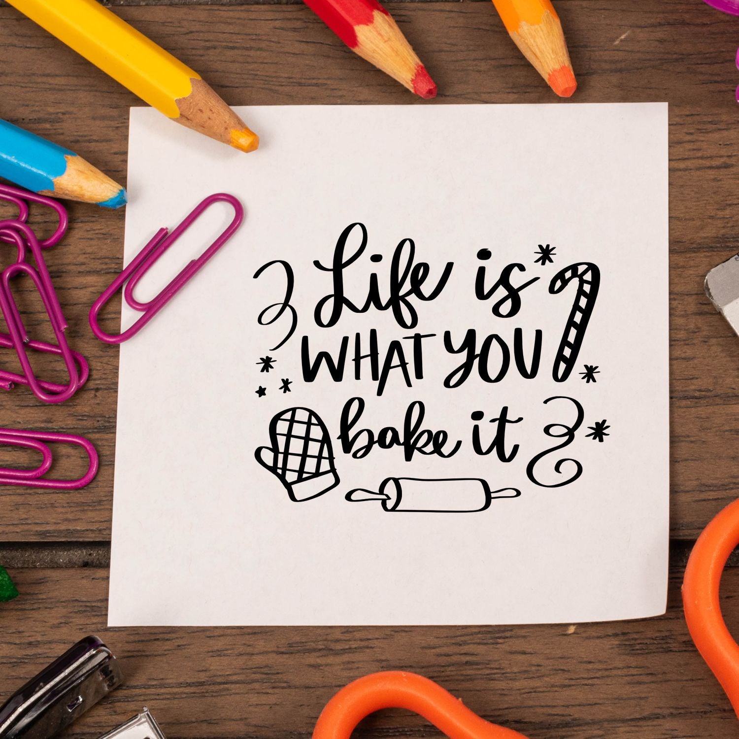 Black stamp and seal design on white paper reading Life is what you bake it, with oven mitt, rolling pin, and colorful pencils.