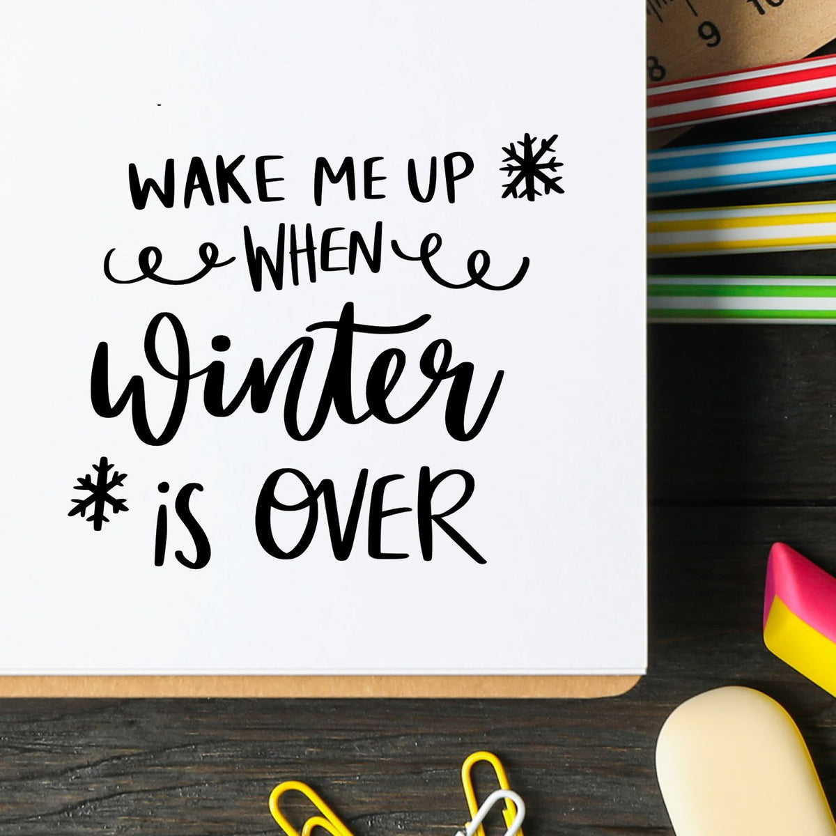 Black winter phrase stamp on white paper with snowflakes, beside colorful stamp and seal supplies on a dark desk.