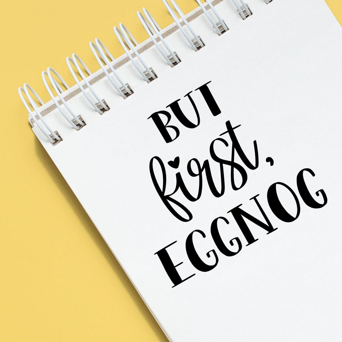 Black stamp and seal design reading But first, eggnog on a white spiral notebook over a yellow background.