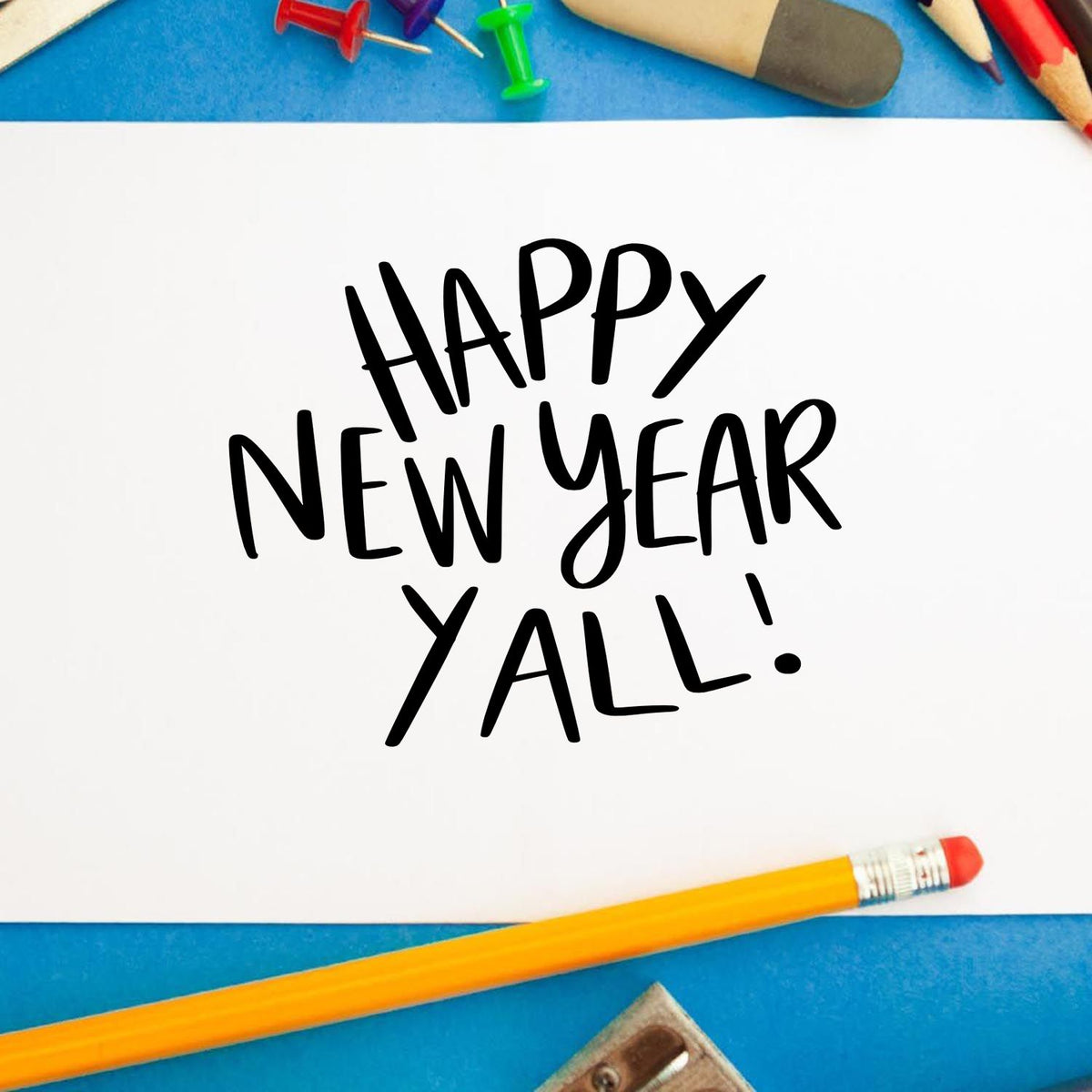Black Happy New Year Y'all! stamp on white paper, framed by blue background and colorful pencils, pins, and sharpener.
