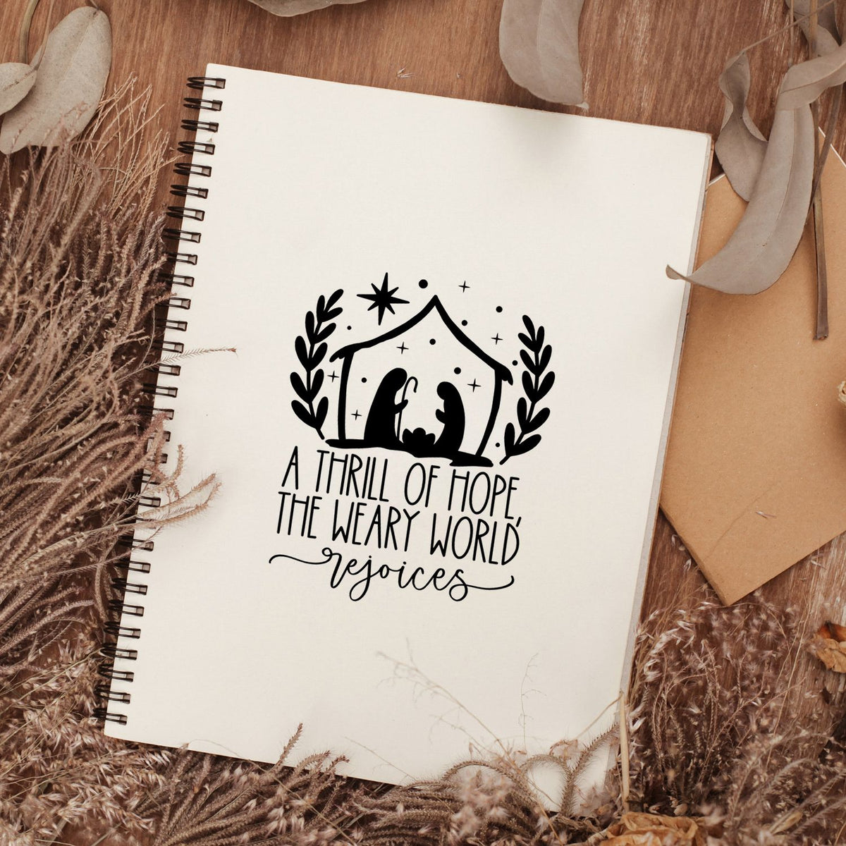 Black stamp seal on a cream spiral notebook, showing a nativity under a star with laurel leaves and A thrill of hope...rejoices.