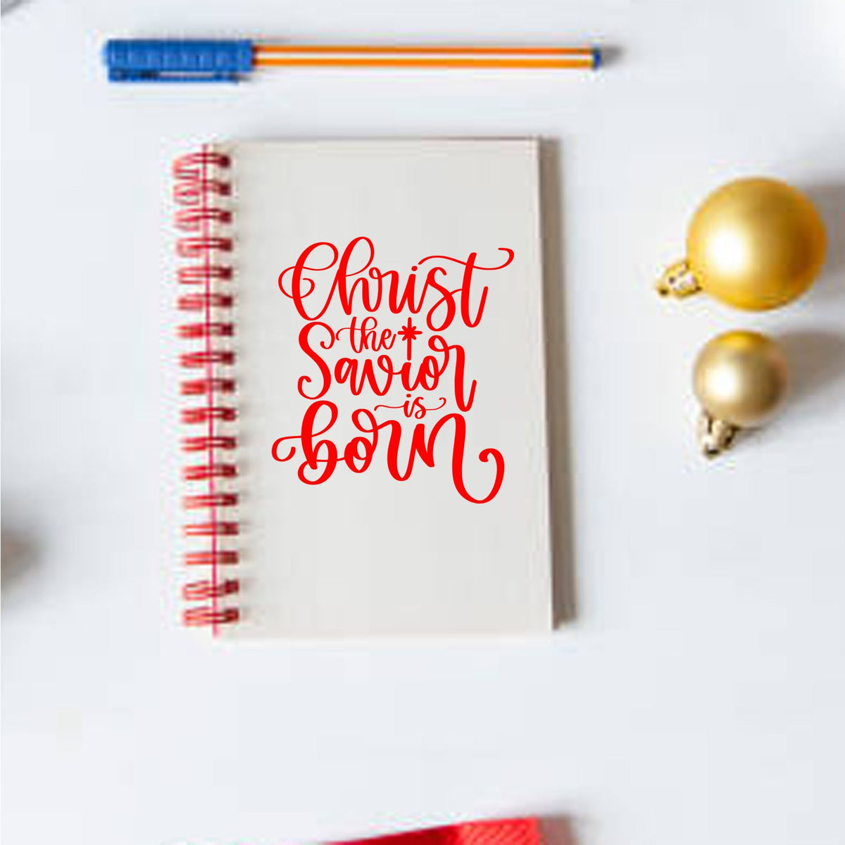 Red holiday stamp reading Christ the Savior is Born on a white spiral notebook, with red binding and gold ornaments.