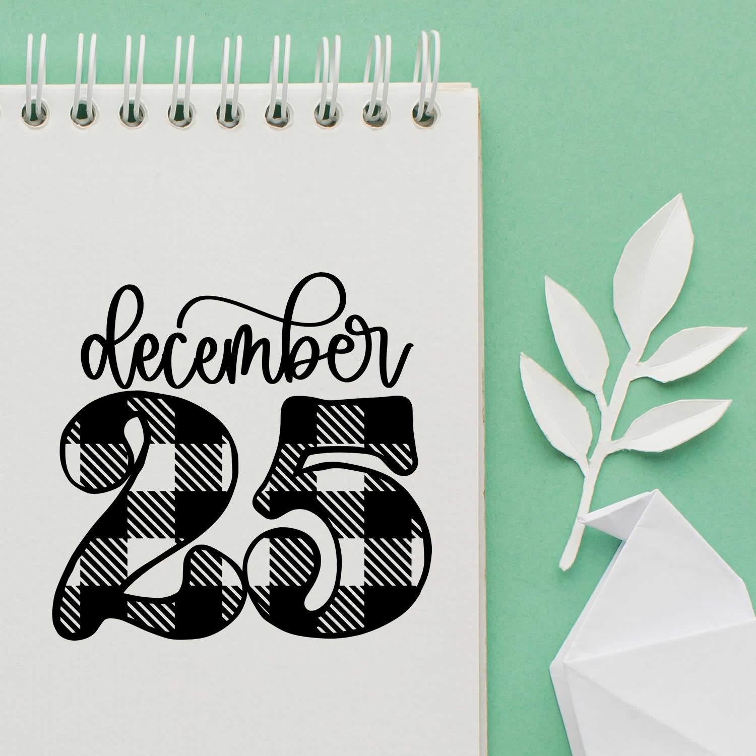 Black December 25 stamp and seal design on white spiral notebook page, with mint green background and white paper leaves.