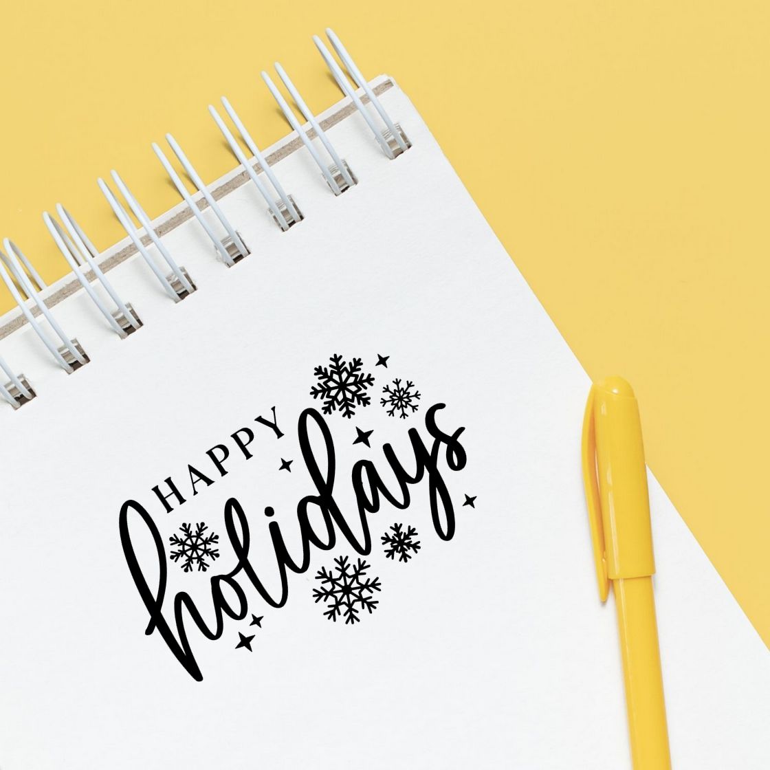 Black Happy Holidays stamp and seal design with snowflakes on a white spiral notebook, beside a yellow pen on a yellow background.