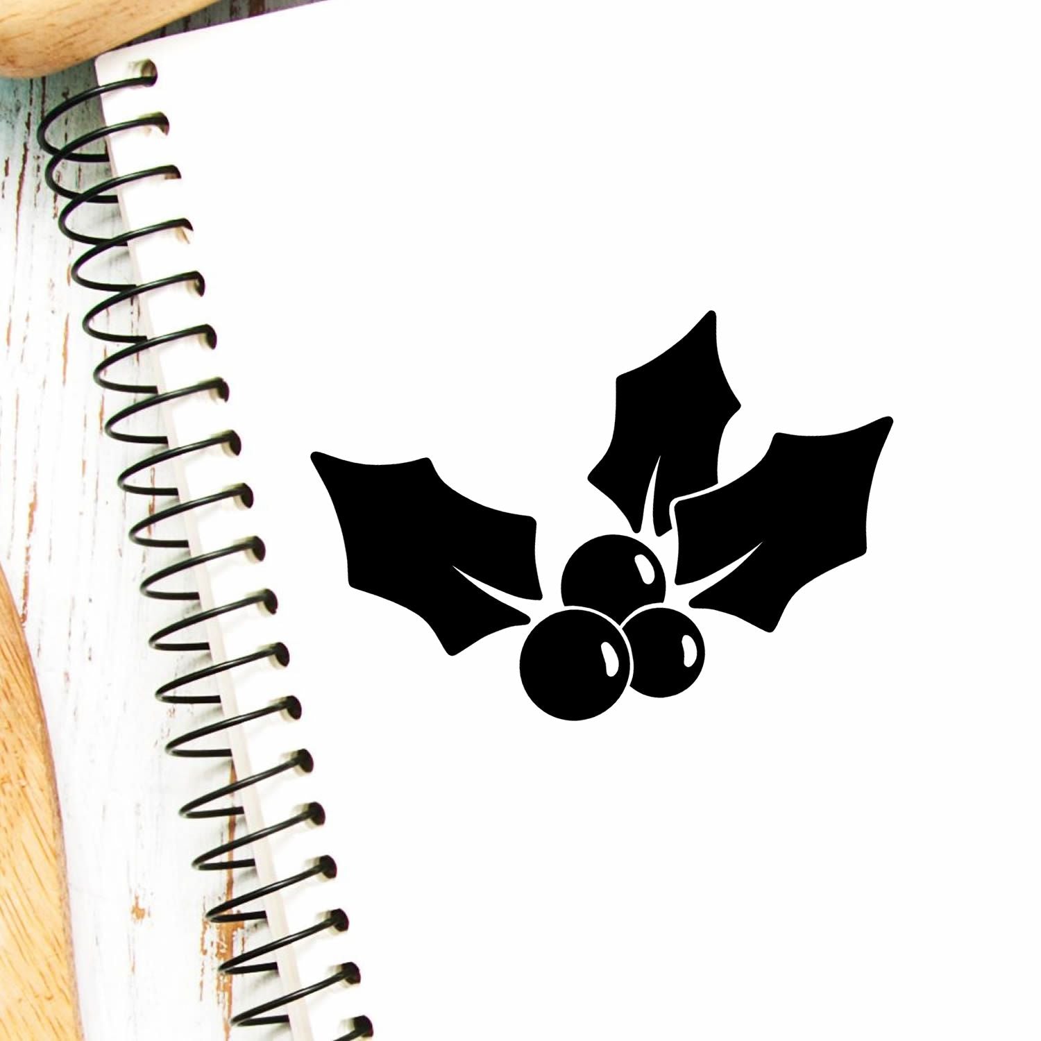 Black holly berry stamp and seal design on white background with spiral notebook edge and rustic wood accents.