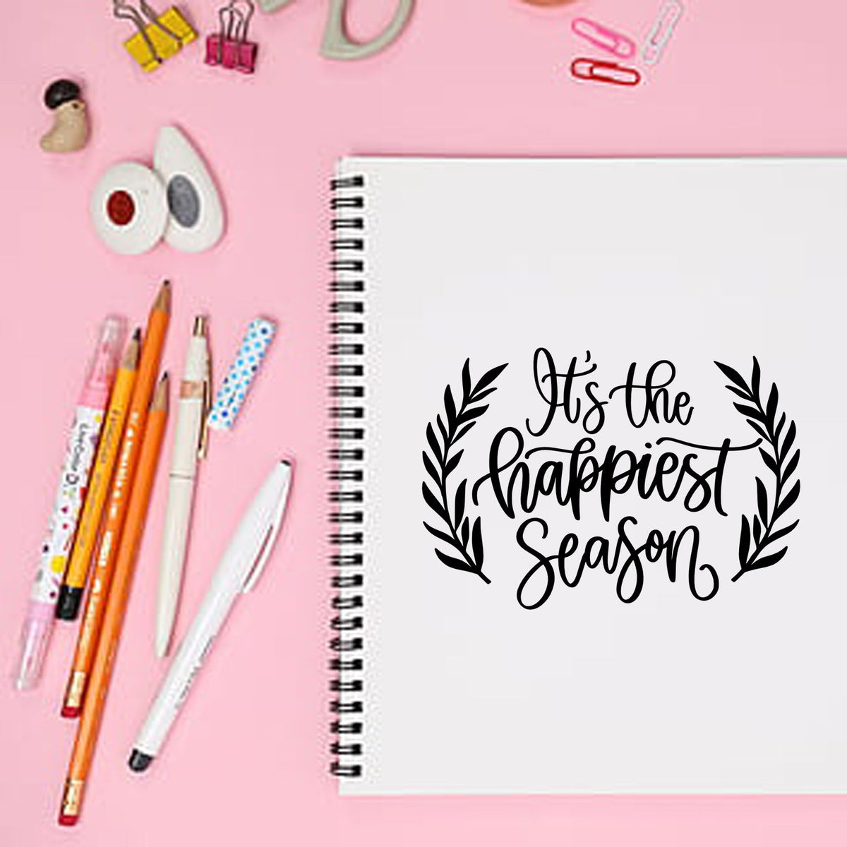 Black holiday stamp seal reading Its the happiest season on white spiral notebook with pink background and stationery.
