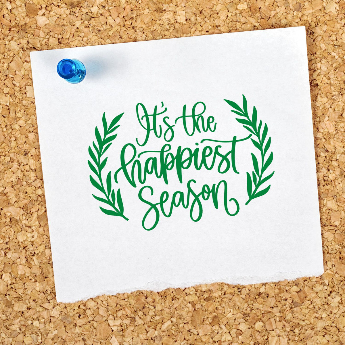 Green stamp and seal with leafy wreath and blue tack on white paper reading Its the happiest season.