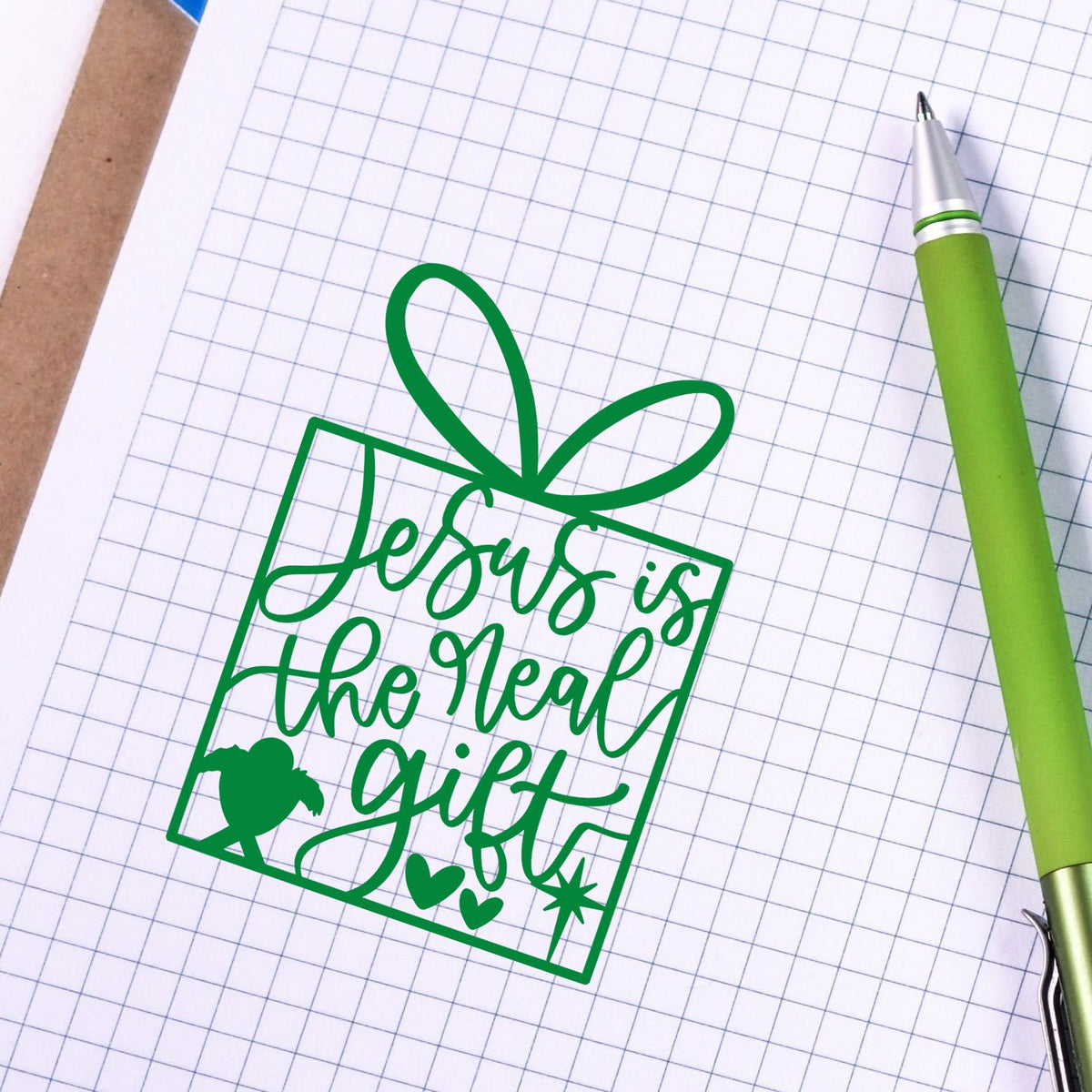 Green stamp seal reading Jesus is the real gift with bow, hearts and star on grid paper, beside a lime green pen.