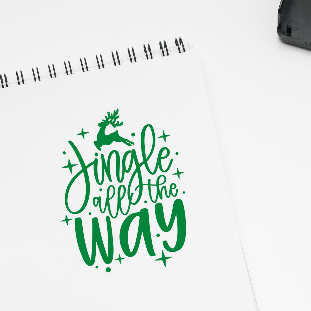 Green holiday stamp seal reading Jingle all the way with deer and stars on white spiral-bound paper.