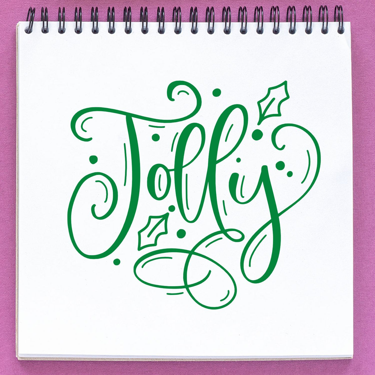 Green Jolly stamp and seal design with festive swirls and holly on a white spiral notebook, pink background.